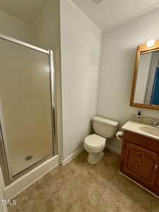 A bathroom with a glass-enclosed shower on the left, a white toilet in the center, and a wooden vanity with a sink, mirror, and light above on the right. The floor is tiled and the walls are white.