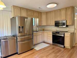 A modern kitchen with light wood cabinets, stainless steel refrigerator, electric stove with oven, built-in microwave, and a sink under a window. The floor is light wood, and a small mat is placed in front of the sink.