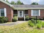 A single-story brick house with white trim at 65 Opie Frazier Road, Henderson. Steps lead to a metal screen door, with shrubs along the front, a lamp post, green plants in the yard, and a concrete walkway curving toward the porch.