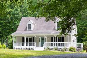 A light-colored, two-story house with a gable roof, front porch, white railing, and blue front door, surrounded by trees and greenery with a gravel driveway in front.