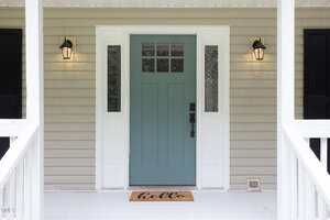 A light green front door with glass panes is centered between two white-framed windows on a beige house. Two black lantern-style lights are mounted on either side, and a doormat with "hello" is in front of the door.