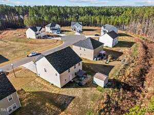 Aerial view of a suburban cul-de-sac with multiple two-story houses, driveways, parked cars, a small shed, fenced backyards, and a wooded area bordering the neighborhood.