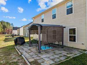 A backyard patio with a pergola over outdoor furniture, a rug, and a covered grill, next to a tan house with several windows; neighboring houses and trees are visible in the background.