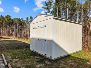A light-colored wooden storage shed with double doors stands on a patch of grass, surrounded by tall trees and under a partly cloudy sky. Shadows from the trees fall across the shed and ground.
