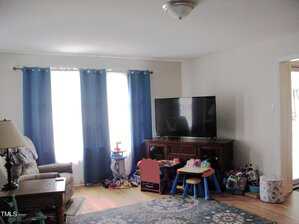 A living room with light walls, blue curtains, a large TV on a wooden stand, children's toys and furniture, an armchair, a side table with a lamp, and a patterned rug on a hardwood floor.
