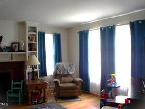 A living room with hardwood floors, blue curtains, three windows, an armchair with a cover, side tables, shelves, a floor lamp, and children's toys near the windows.