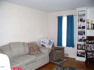 A living room with a beige sofa, patterned pillows, a brown recliner with a blanket, blue curtains on a window, hardwood floor, and built-in shelves holding books, photos, and decorative items.
