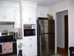 A kitchen with white cabinets, a stainless steel refrigerator, a microwave in a built-in shelf, a black stove with a colorful towel, and various items on top of the fridge and counter.