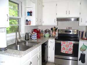 A kitchen with white cabinets, a marble-patterned countertop, an electric stove with a floral towel, a double sink, various utensils, small appliances, and a window with a view of green trees.