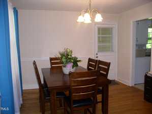 A dining room with a dark wooden table and six matching chairs, a floral centerpiece on the table, hardwood floors, a hanging light fixture, a door with a window, and an open doorway leading to a kitchen.