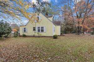 A yellow two-story house with white trim sits on a grassy yard scattered with autumn leaves. Several windows are visible on the side of the house, and trees with fall foliage surround the property.