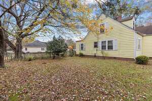 A yellow house with white trim sits behind a yard covered in fallen leaves. A large tree with yellow leaves stands near a wooden picket fence. There are bushes along the house and a neighboring house is visible in the background.