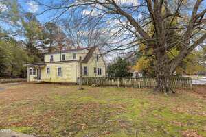 A two-story yellow house with a sloped roof and white trim sits behind a white picket fence. Leafless trees and sparse grass with patches of moss cover the yard under a partly cloudy sky.