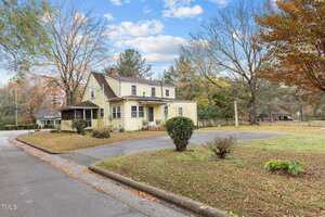 A two-story yellow house with white trim sits on a corner lot with a paved driveway, some small shrubs, and trees with autumn foliage. The sky is partly cloudy.