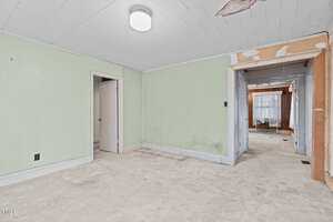 A mostly empty room with light green walls, white trim, and a worn, stained carpet. The ceiling panels are white, with some damage near a light fixture. There are doorways leading to other rooms.