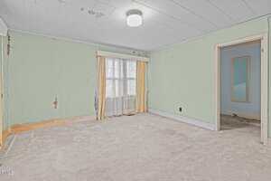 An empty room with pale green walls, some peeling paint, a window with sheer curtains and yellow drapes, beige carpet, and an open doorway leading to another room with blue walls.