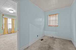 A small empty room with light blue walls, a single window with sheer peach curtains, and a beige carpet. An open doorway leads to a larger room with pale green walls and yellow curtains.