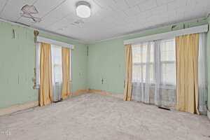 Empty room with pale green walls, two windows with yellow curtains, a white ceiling light, and visible water damage on the ceiling and walls. The floor is carpeted in a light color.