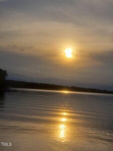 The sun is setting over a calm lake, casting a golden reflection on the water. The sky is mostly cloudy with some visible rays of sunlight. Trees and a dock are faintly visible along the shoreline.