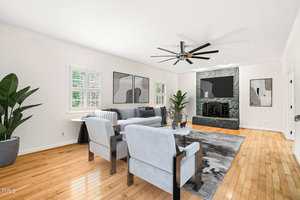 A modern living room with light hardwood floors, white walls, gray furniture, abstract wall art, a large television, two potted plants, and a ceiling fan. Natural light enters through shuttered windows.