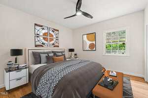 A modern bedroom with a double bed featuring gray and white bedding, two white nightstands with lamps, a brown ottoman, wall art, a ceiling fan, and a window showing trees and a fenced yard outside.
