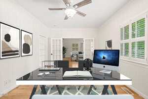 A modern home office with a black and white desk, an iMac, open notebook, Abacus, and headphones. Artwork hangs on the wall, and French doors open to another room with a sofa and plants. White shutters cover the windows.