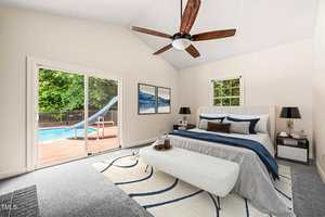 A bedroom with a bed, white bench, two nightstands, wall art, and a ceiling fan. A large window and sliding glass door open to a patio with a swimming pool and slide. The room has light walls and a patterned rug.