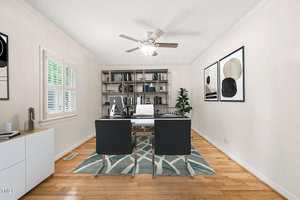 A home office with two black chairs facing a desk, a white chair behind the desk, bookshelves against the back wall, abstract art on the walls, a ceiling fan, a potted plant, and a window with white shutters.