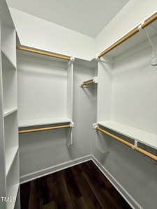 A walk-in closet with white shelves and hanging rods on three walls, featuring dark wood flooring and white walls. The closet includes multiple storage sections and open shelving.