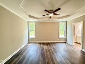 Empty room with beige walls, dark wood flooring, a ceiling fan with lights, two windows on either side of the wall, and a doorway leading to a bathroom with a visible vanity and sink.