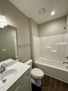 A small bathroom with a white vanity and sink, a mirror above, a toilet, and a shower-tub combo with a built-in shelf. The walls are beige, and the floor is dark wood or wood-look material.