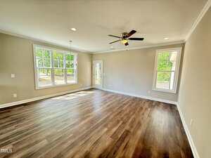 Bright, empty living room with beige walls, wood flooring, large windows, a ceiling fan with lights, and a glass door leading outside. Natural light fills the space and trees are visible through the windows.