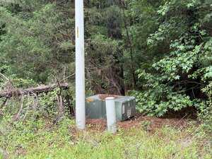 A green utility box and two gray utility posts sit on the ground near a tall utility pole, surrounded by dense trees and vegetation. A fallen tree trunk lies nearby.