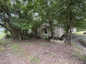A small, weathered wooden house sits surrounded by overgrown trees and brush at 00 Warrenton Road. The house has peeling paint, a missing step at the entrance, and an empty, rectangular concrete planter in front.