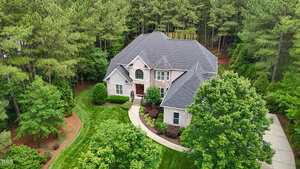A two-story brick house with a dark gray roof is surrounded by green trees and landscaped lawns, with a curved walkway leading to the front entrance and a driveway on the right.