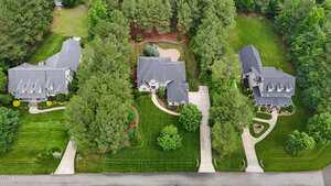 Aerial view of three large suburban houses with grey roofs, surrounded by green lawns, trees, and landscaped gardens, each with a driveway leading to the street at the bottom of the image.