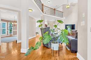 A living room with hardwood floors, a large potted monstera plant in the foreground, a gray armchair, a black sofa, a TV on a wooden stand, and a staircase with a decorative railing in the background.