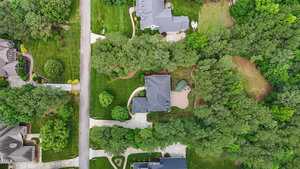 Aerial view of a suburban neighborhood with large houses, driveways, manicured lawns, and surrounding trees. Several homes are separated by roads and patches of dense greenery.