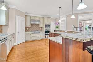 A spacious kitchen with wooden floors, beige cabinets, stainless steel appliances, granite countertops, pendant lights, and a central island with a raised bar area. Purple flowers decorate the countertop.