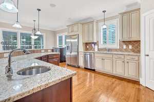 Spacious kitchen with granite countertops, stainless steel appliances, white cabinets, a center island with a sink, pendant lighting, tiled backsplash, and hardwood floors. Large windows provide natural light.
