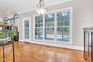 A dining area with hardwood floors, a hanging light fixture, large windows, a glass door, and views of a screened porch and trees outside. A dark cabinet stands against the right wall.