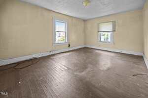Empty room with beige walls, two windows with white trim, brown wooden floor, some scuff marks and stains on the walls and floor, a ceiling light fixture, and visible electrical cords along the baseboard.
