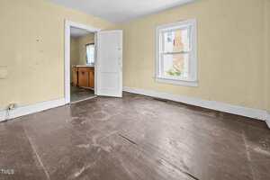 Empty room with worn brown wood floors, yellow walls, white trim, a single window, and an open door leading to a small kitchen area with wood cabinets and visible countertop.