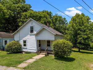 A small white house with an A-frame roof, front porch, and steps leading to the entrance. Two large bushes flank the porch, with grass and trees surrounding the home under a partly cloudy sky.