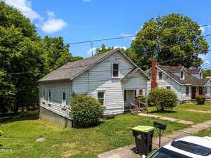 A white, single-story house with an attic and gray roof sits on a grassy lot. There are bushes near the front steps, overhead power lines, a driveway, and a green garbage bin at the curb. Neighboring houses are visible.