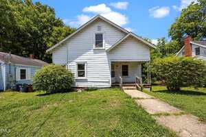 A white, two-story house with a sloped roof and small front porch, surrounded by grass, bushes, and trees. A concrete path leads from the sidewalk to the front door. The sky is blue with a few clouds.