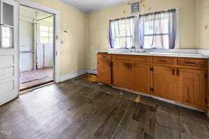 An old kitchen with worn wooden cabinets, damaged flooring, and two curtained windows above the counter. An open doorway leads to a sunlit, unfinished adjoining room. The walls are cream-colored.