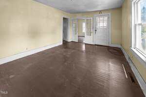An empty room with brown painted wooden floors, pale yellow walls, two large windows on the right, and glass-paneled doors leading to another room. There is visible wear on the floor and some marks on the walls.