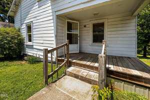 A small wooden porch with a railing leads to the front door of a white house with horizontal siding. There is a window on the left and a shrub next to the porch. The grass lawn is visible.