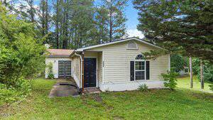 Single-story beige house at 2396 Spring Valley Road, Henderson, with black doors and shutters, set on a grassy lawn with trees in the background. Features a gabled roof and a concrete walkway leading to the entrance.